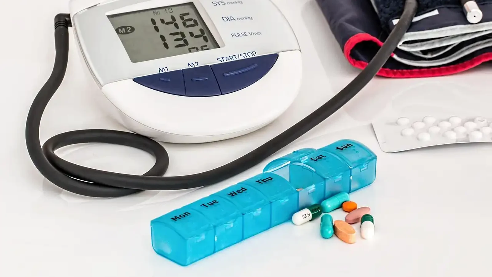 A digital upper-arm blood pressure monitor showing a reading, its tube coiled beside a folded cuff, with a blue Monday-to-Sunday pill organiser, loose tablets and capsules, and a blister pack in front — what do blood pressure numbers mean