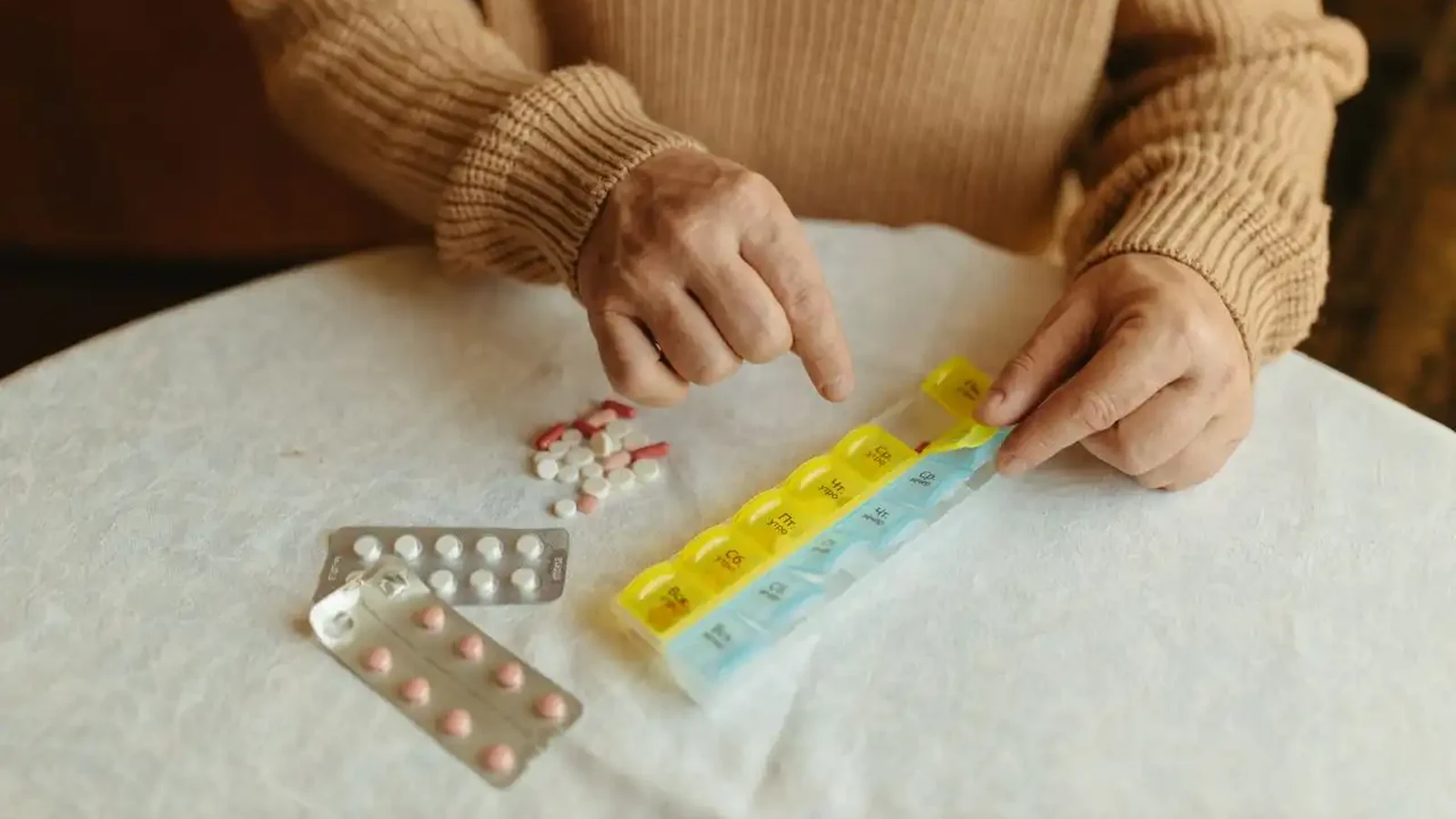 Older person in a beige knitted jumper sorting tablets from blister packs into a yellow and blue weekly pill organiser on a white tablecloth — what services can a pharmacy provide